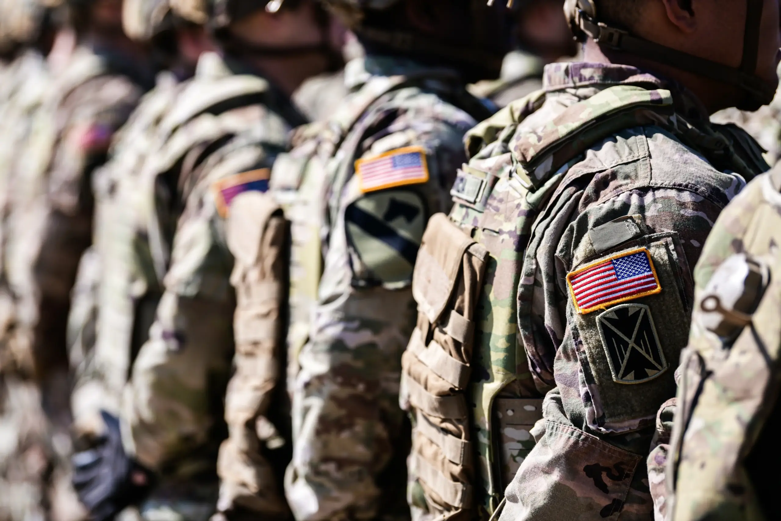 A row of soldiers in camouflage uniforms stand in formation, each with an American flag patch on their right shoulder. Their faces are out of focus or not visible. The scene conveys a sense of unity and military discipline.