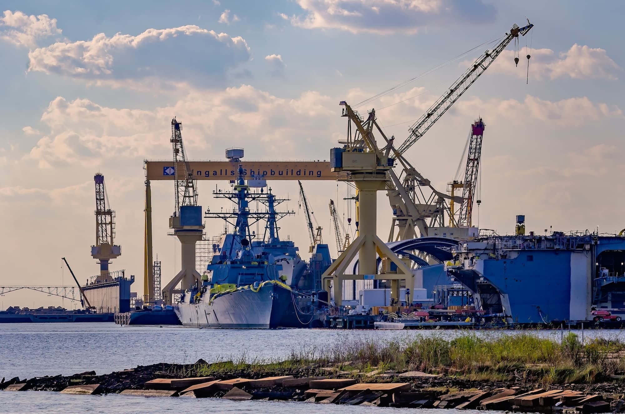 A shipyard with large cranes and a navy ship docked beside an Ingalls Shipbuilding facility; blue sky with scattered clouds is visible in the background.