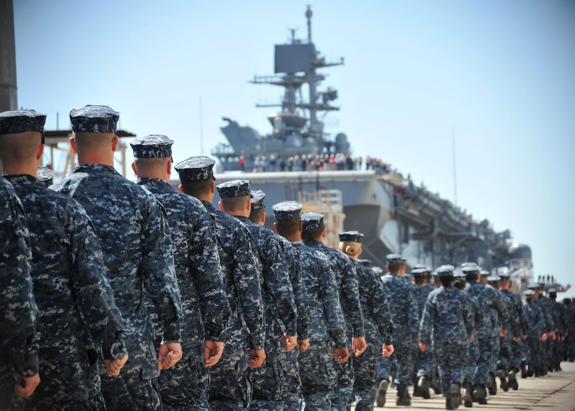 Rows of uniformed Navy sailors walk in formation toward a large aircraft carrier docked at a harbor, with sailors lined up on the deck under a clear blue sky.