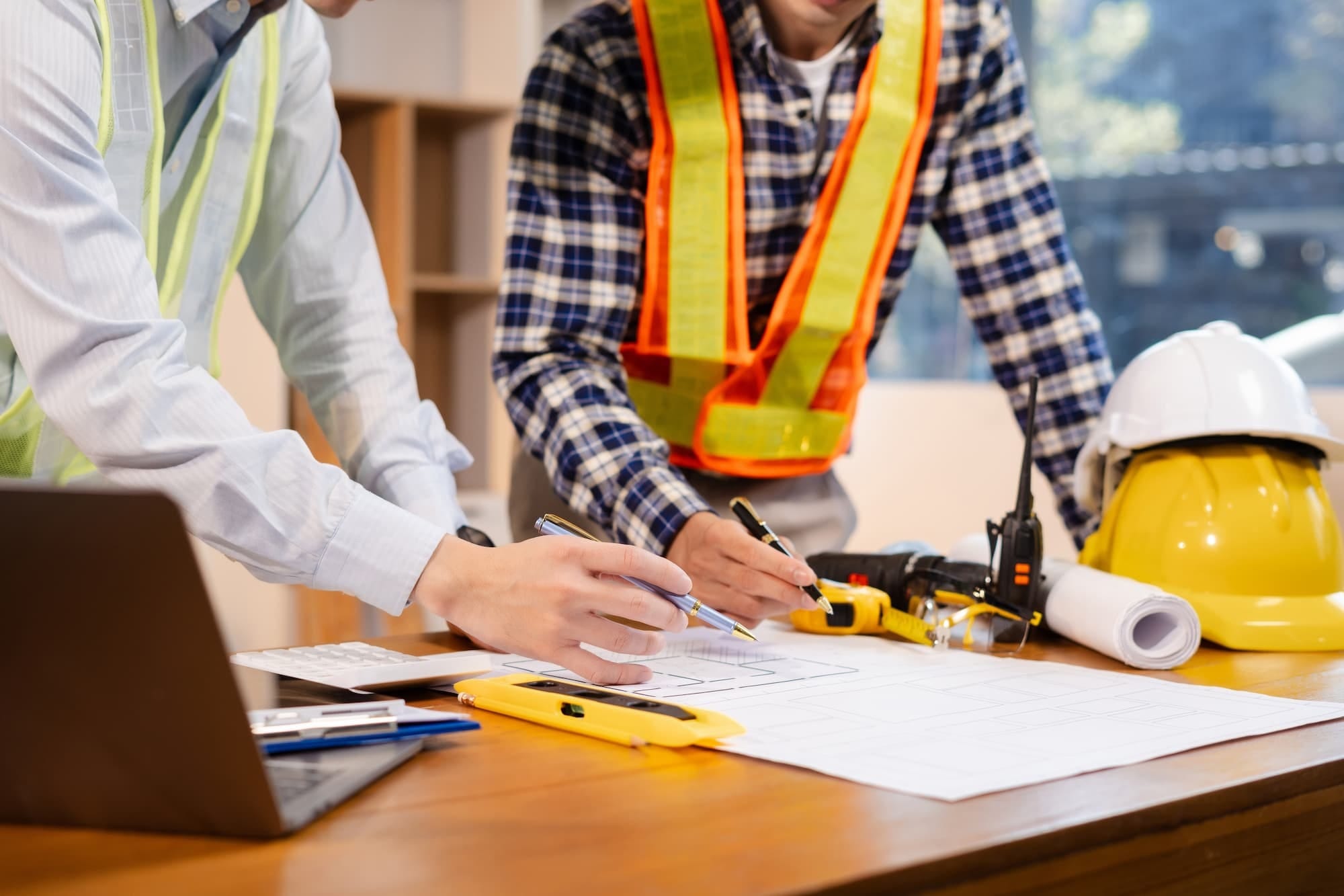 Two people wearing safety vests review blueprints on a desk with a laptop, ruler, walkie-talkie, measuring tape, and safety helmets placed nearby.