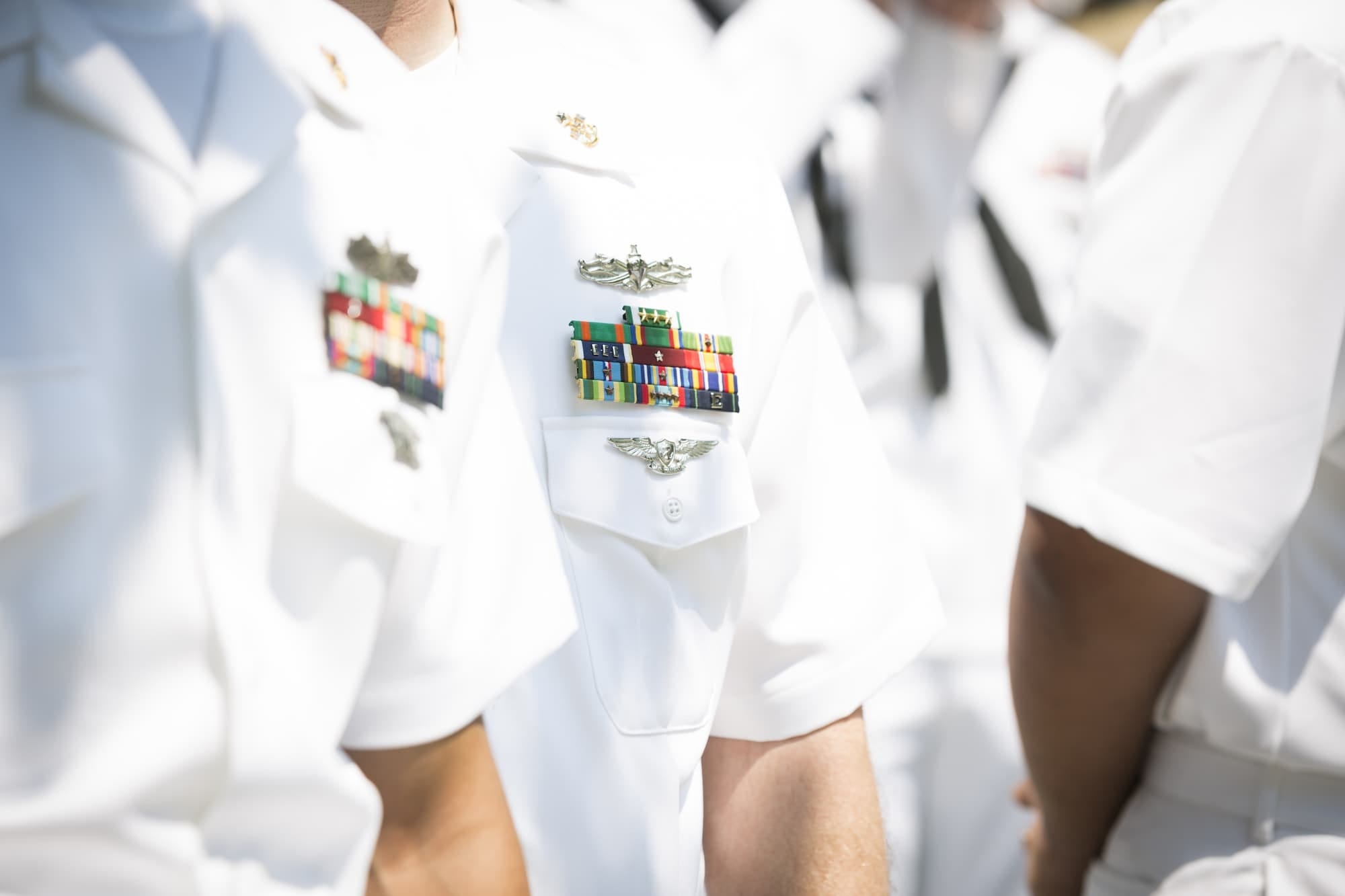Close-up of U.S. Navy personnel in white uniforms with colorful service ribbons and badges on their chests, standing in formation outdoors.