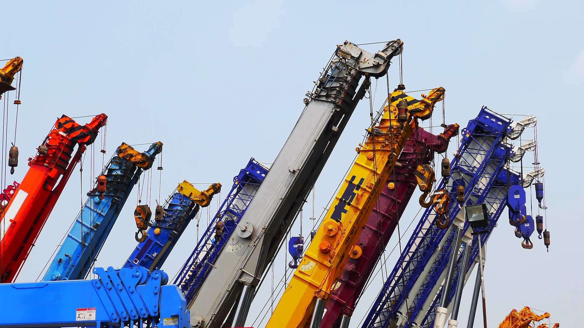 Several colorful construction cranes with their arms raised are lined up against a clear blue sky, showcasing various shades including blue, yellow, red, purple, and gray.