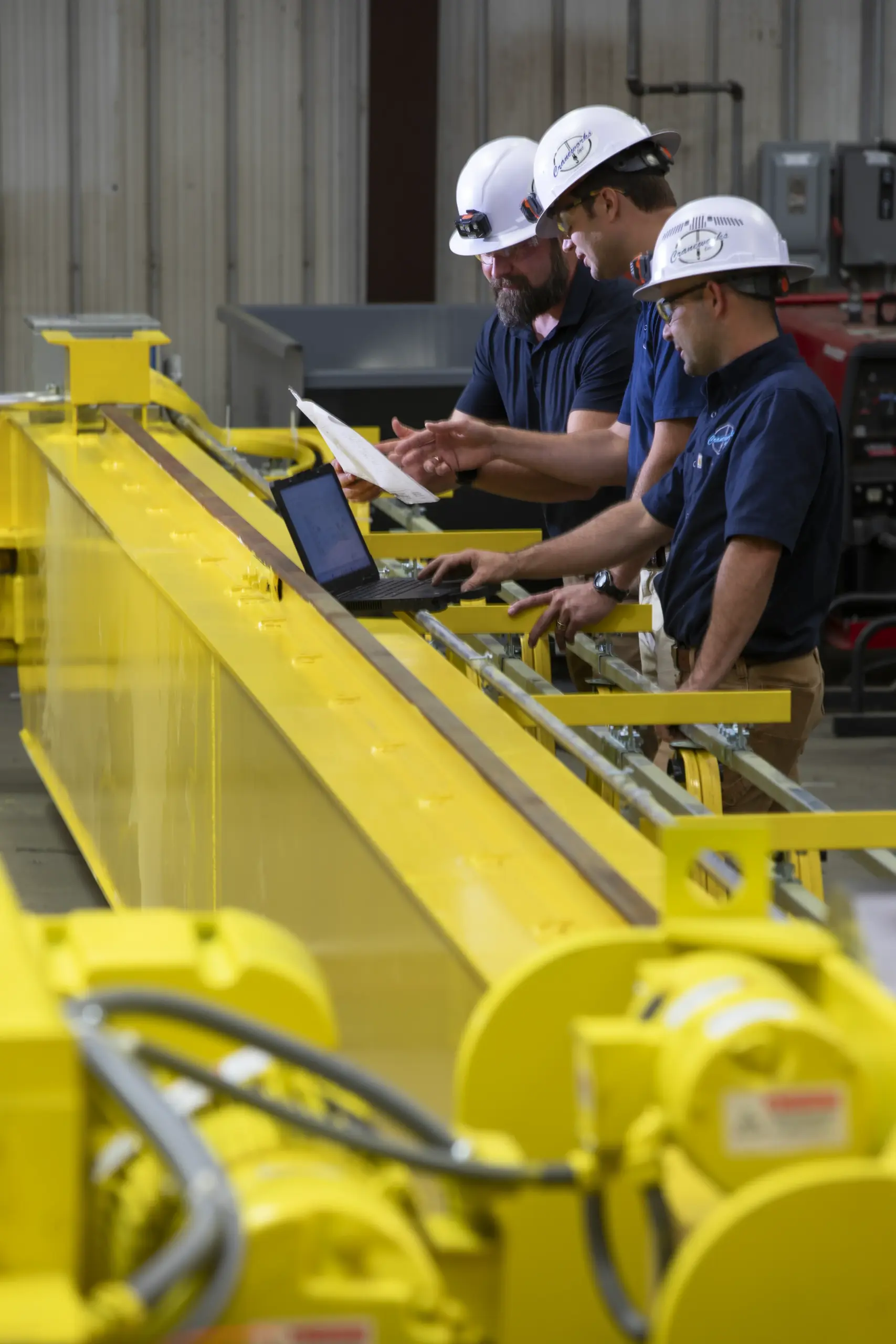 Three men wearing hard hats and navy shirts work together in an industrial setting, examining documents and using a laptop on a long yellow piece of machinery inside a warehouse.